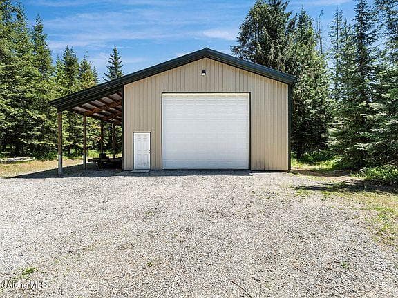Tan metal workshop with a white garage door and lean-to, surrounded by evergreen trees.