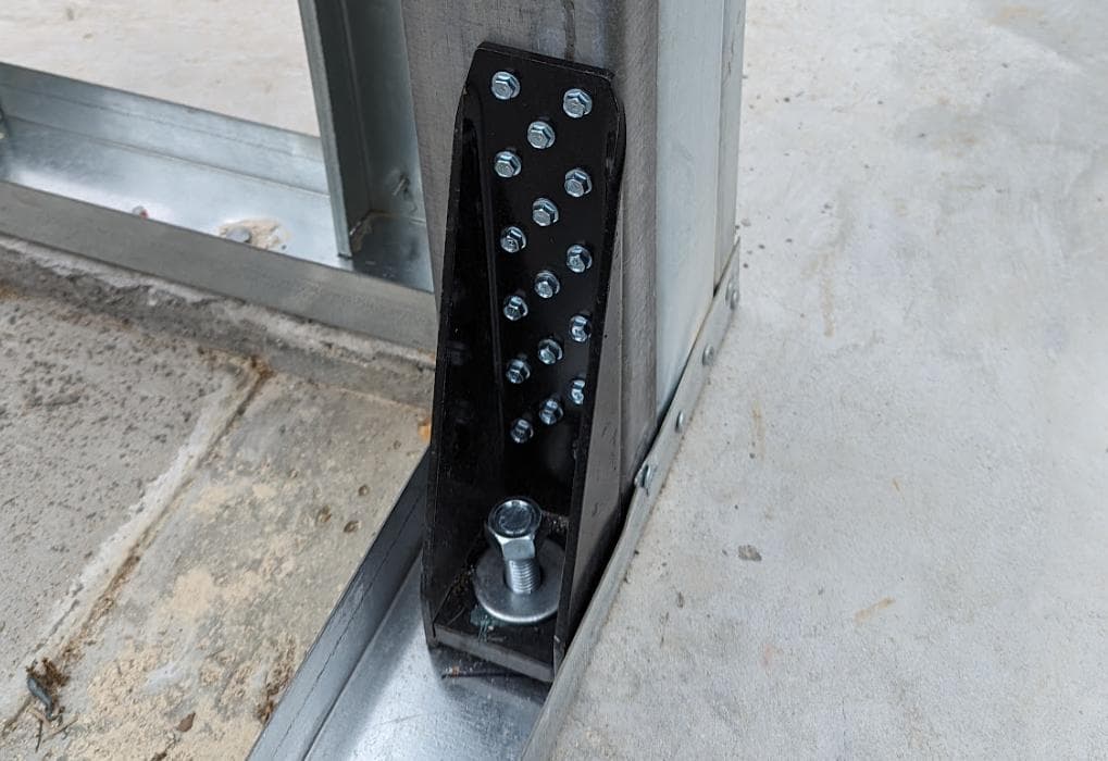 Black metal hold-down bracket securing a vertical steel stud to a concrete foundation with bolts.