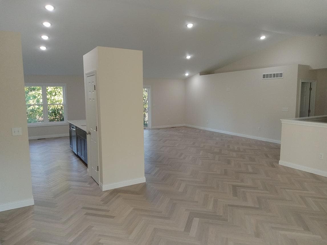 Spacious empty room featuring light-colored herringbone wood floors, white walls, and recessed ceiling lighting.