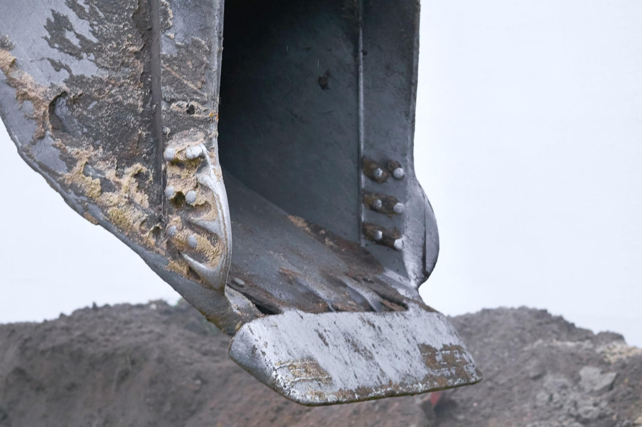 Close-up of a dirty metal excavator bucket hanging over a pile of dark soil.
